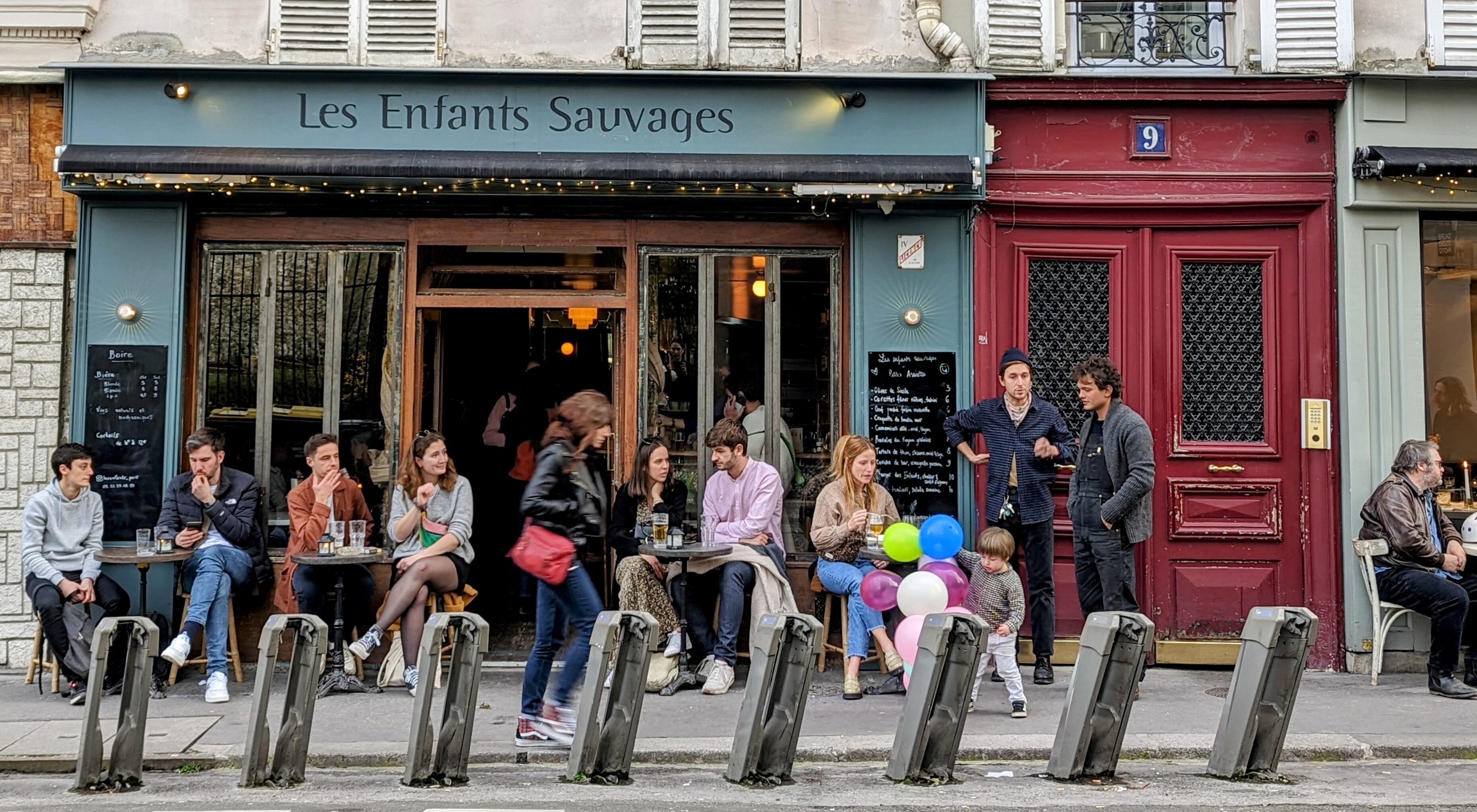 A child playing with balloons and Parisians enjoying drinks on the terrace of a popular neighborhood bistro.